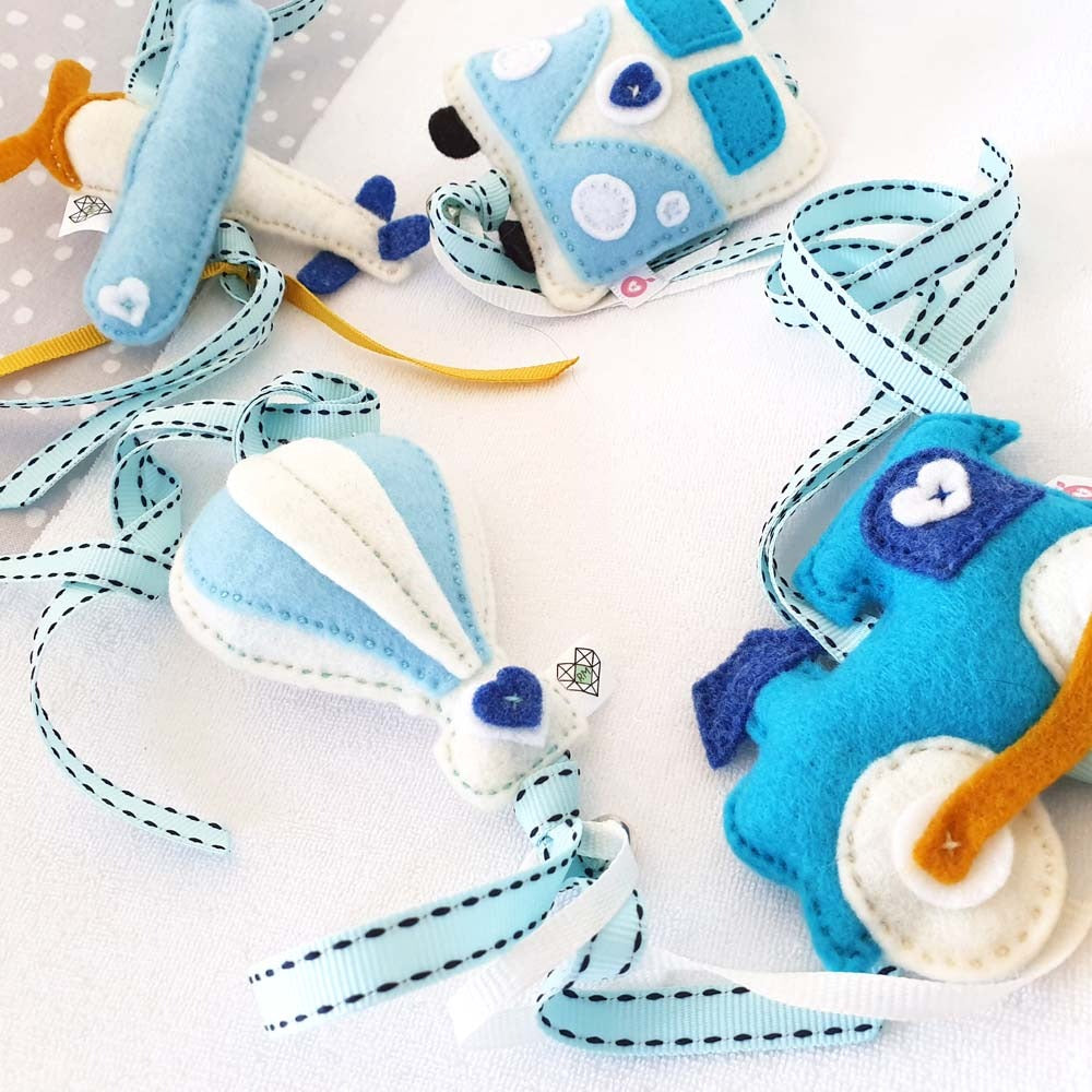 A set of small felt baby accessories, including a hot air balloon, a house, and a dinosaur, crafted in light blue, white, and dark blue, with matching patterned ribbons.