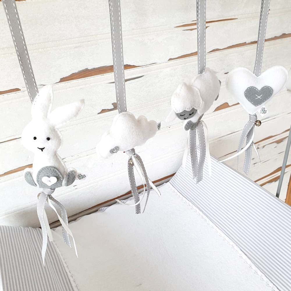 A baby crib hanging toy set featuring a white bunny, fluffy white cloud, sheep, and heart-shaped bird in soft white and gray fabric, suspended by gray ribbons against a distressed white wood crib background.
